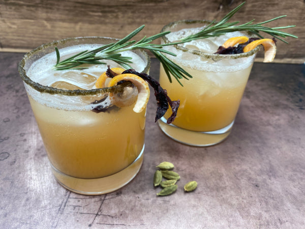 two rocks glasses with frothy grapefruit cocktails filled with ice and garnishde with a spiral of grapefruit rind, a piece of lacer, and a sprig of rosemary. The glass is rimmed with a mixture of brown kelp powder and sugar