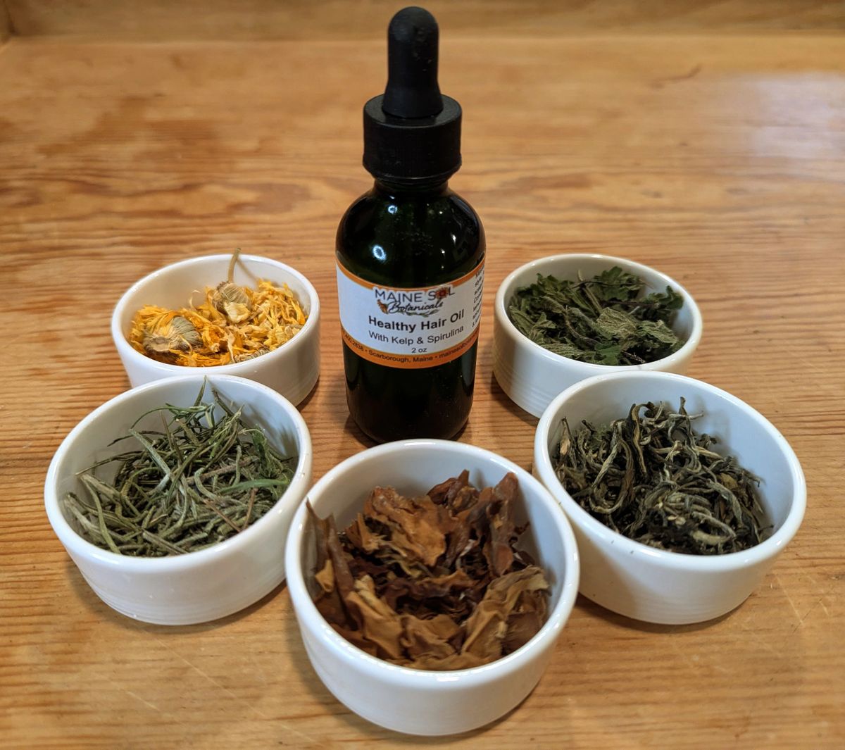 Bottle of 'Healthy Hair Oil' with dried herbs in small white bowls on a wooden surface