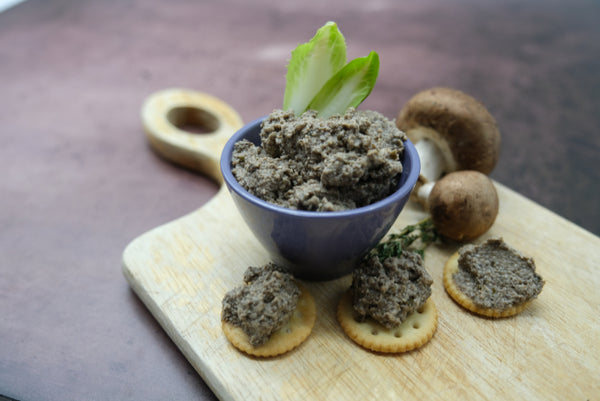 Mushroom Pate in a bowl with three leaves of endive poking out, three crackers with pate on them, two whole mushrooms on a wooden cutting board