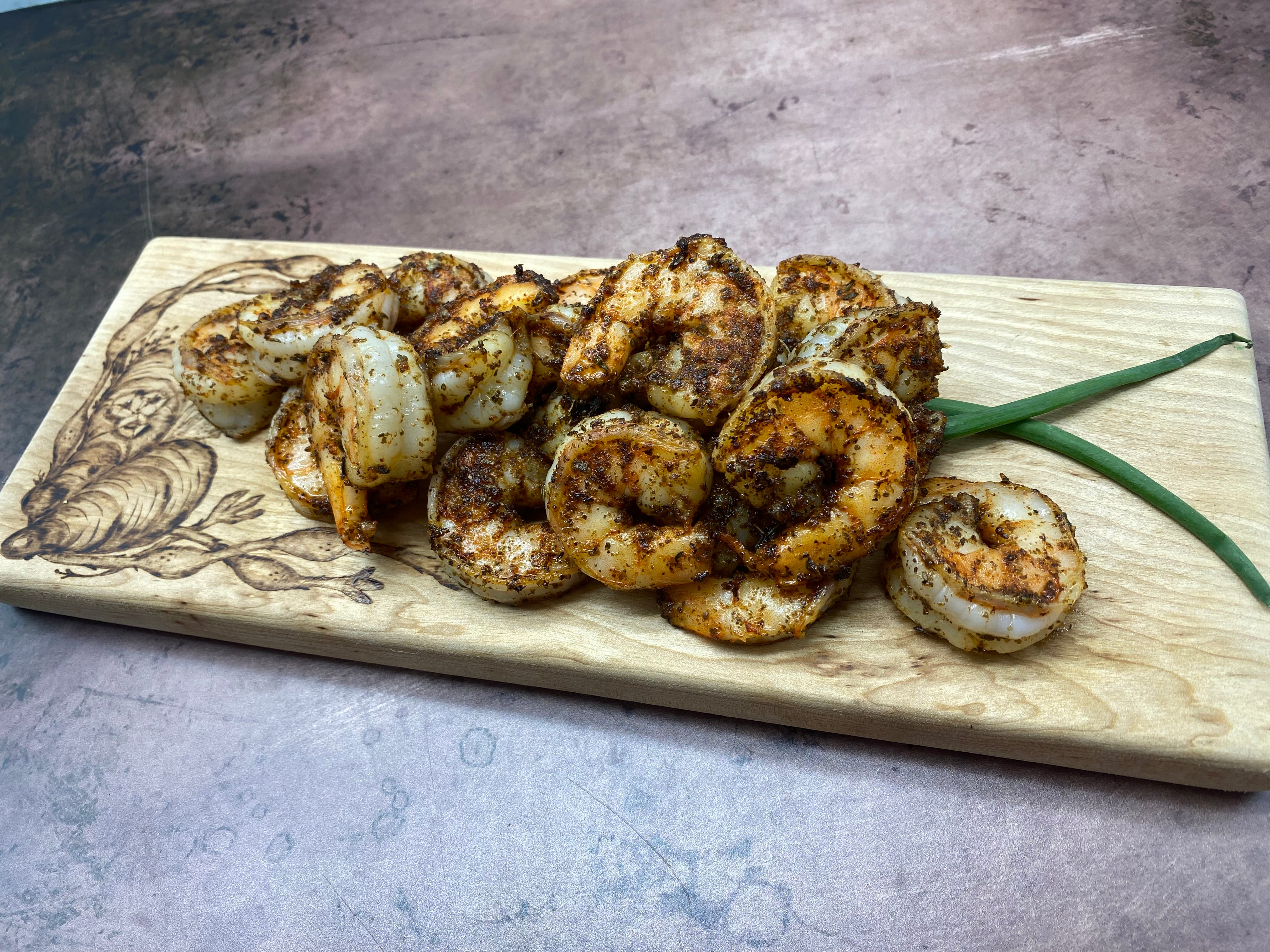 wooden cutting board with wood-burned image of mussels and seaweed piled with browned shrimp coated in dark spices