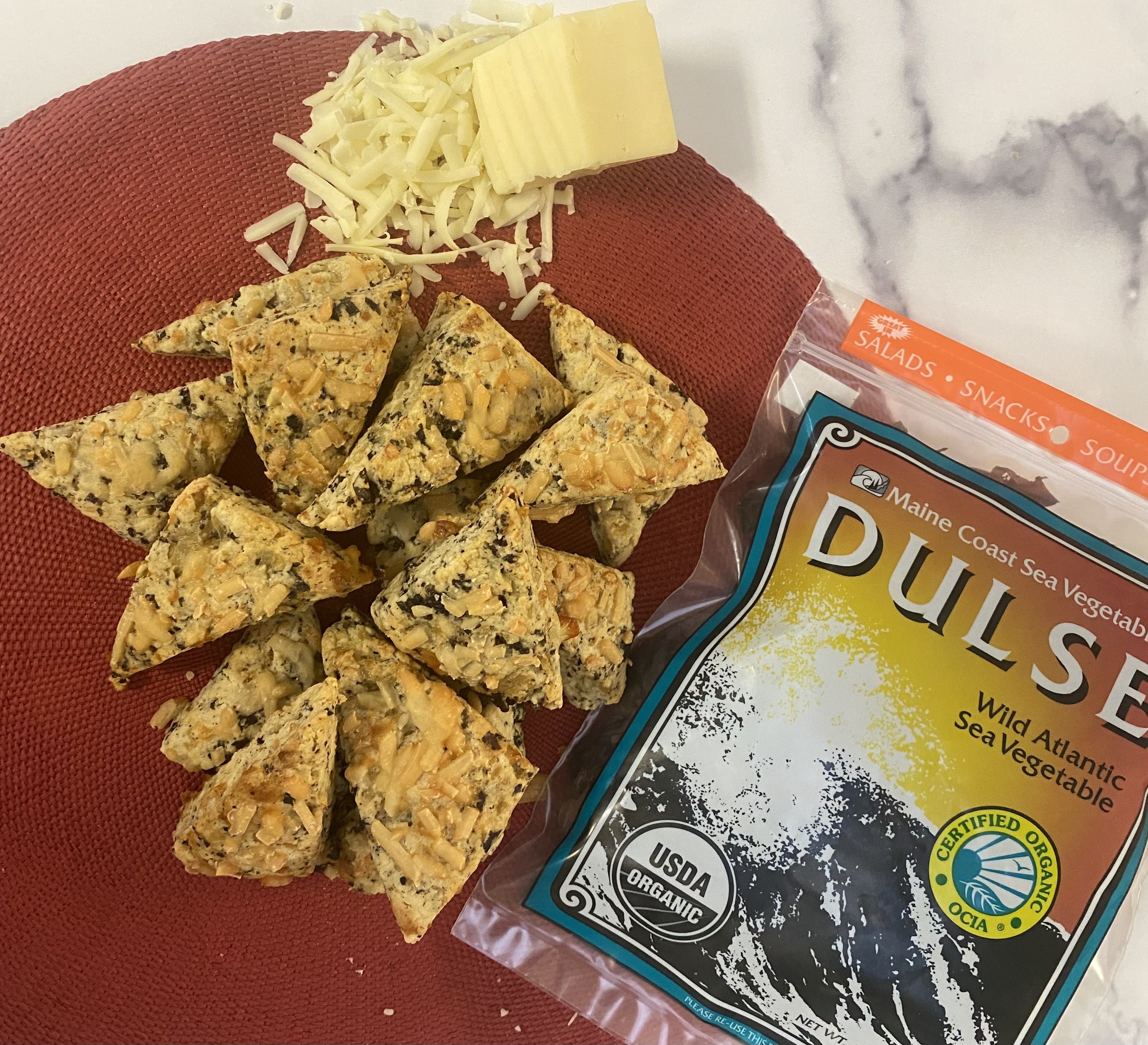 A red placemat with triangle shaped scones flecked with red seaweed. Apile of shredded cheese, a small block of cheese, and a retail package of dulse surround the scones on a white and gray marble background.